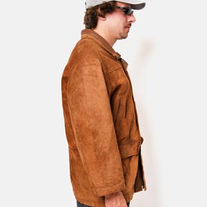 May include: A brown suede jacket with a button closure and a flap pocket on the chest. The jacket has a gray baseball cap on top.