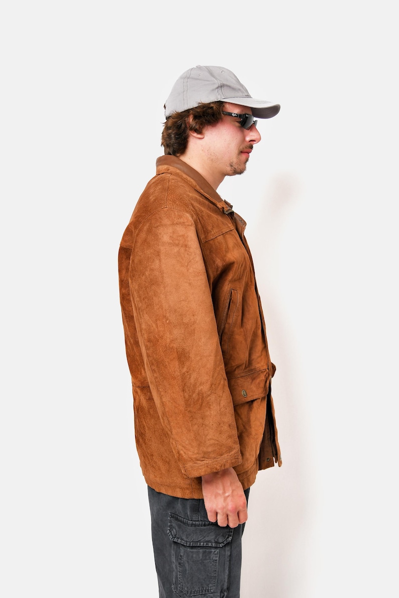 May include: A brown suede jacket with a button closure and a flap pocket on the chest. The jacket has a gray baseball cap on top.