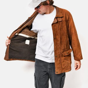 May include: A brown suede jacket with a button closure and a flap pocket on the left side. The jacket is open and the lining is visible. The person is wearing a white t-shirt and gray pants.