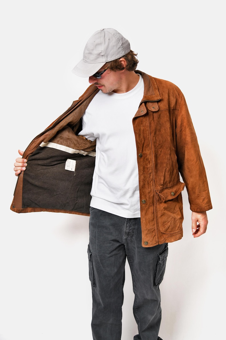 May include: A brown suede jacket with a button closure and a flap pocket on the left side. The jacket is open and the lining is visible. The person is wearing a white t-shirt and gray pants.