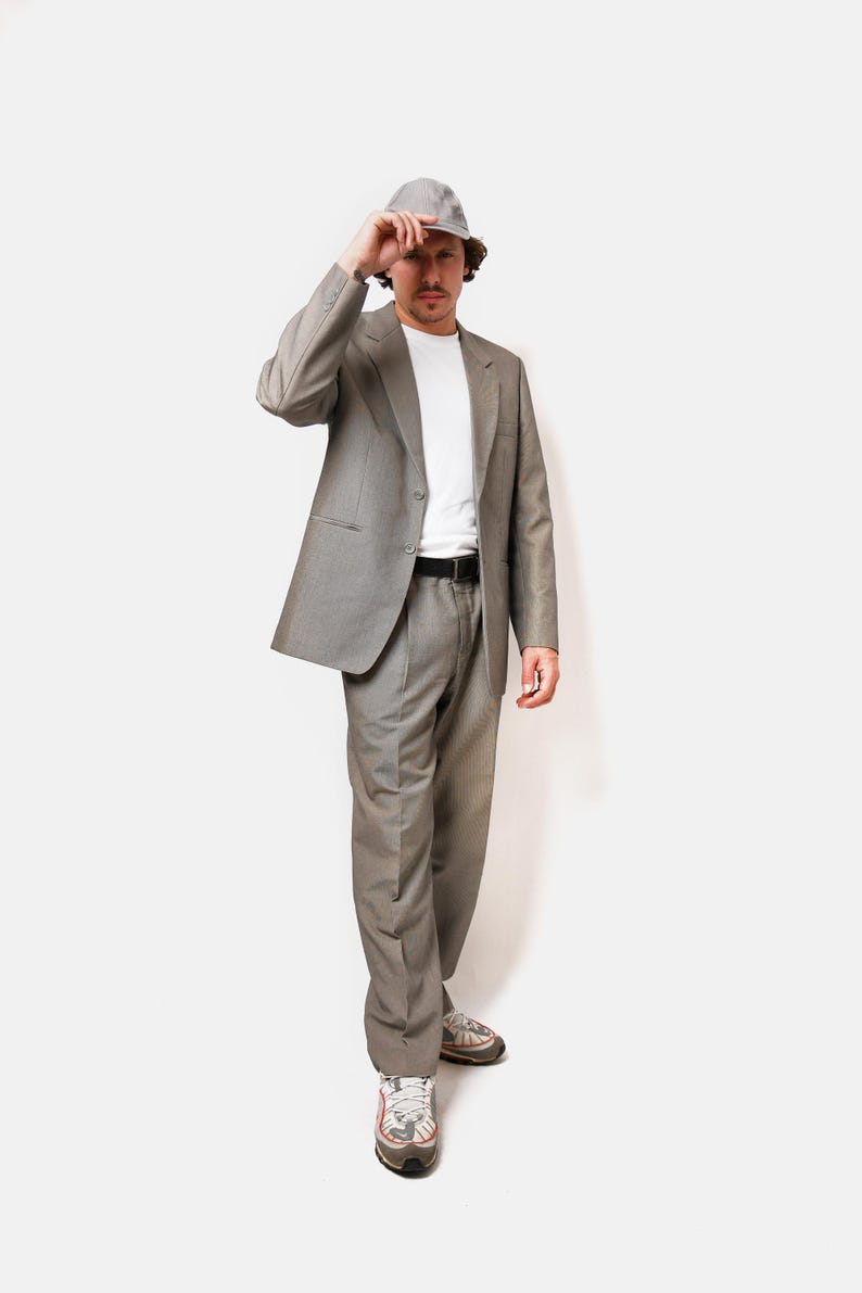 May include: A man wearing a gray suit with a white t-shirt and a gray bucket hat. He is wearing white sneakers with red accents.