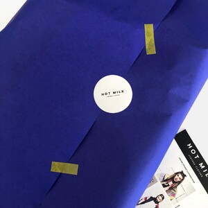 May include: A package wrapped in deep blue paper, sealed with gold tape and a white circular sticker with the text "HOT MILK VINTAGE CLOTHES". A promotional card with the text "HOT MILK ONLINE VINTAGE CLOTHING SHOP" is visible.