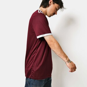 May include: A maroon t-shirt with white trim on the collar and sleeves. The person is wearing blue jeans.