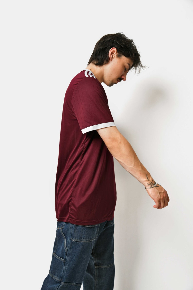 May include: A maroon t-shirt with white trim on the collar and sleeves. The person is wearing blue jeans.