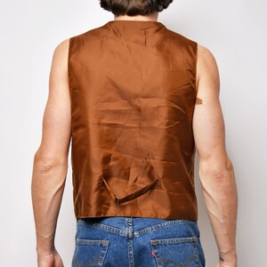 May include: A brown sleeveless vest with a single back strap. The vest is made of a smooth, silky material. The person wearing the vest is also wearing blue jeans and a brown baseball cap with the word "CALIFORNIA" on the back.