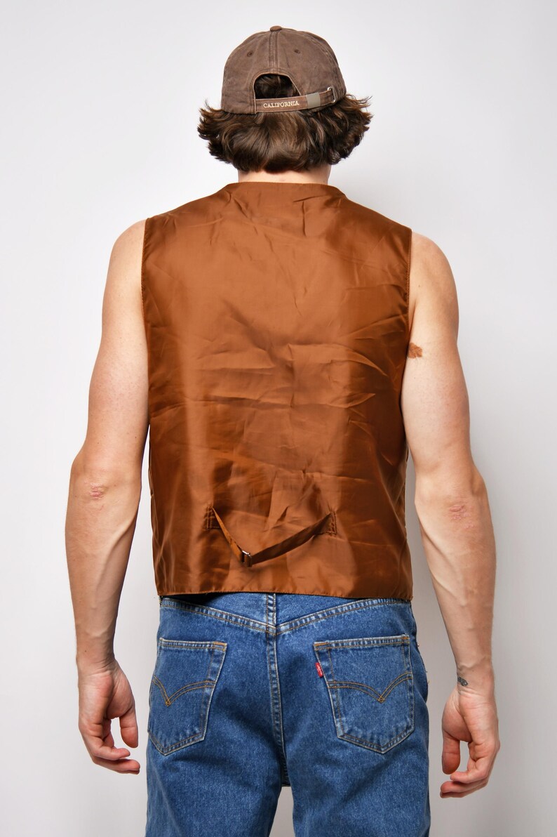 May include: A brown sleeveless vest with a single back strap. The vest is made of a smooth, silky material. The person wearing the vest is also wearing blue jeans and a brown baseball cap with the word "CALIFORNIA" on the back.