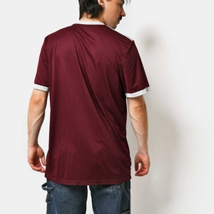 May include: A maroon t-shirt with white trim on the collar and sleeves. The t-shirt is made of a soft, comfortable material and has a relaxed fit. The person is wearing blue jeans.