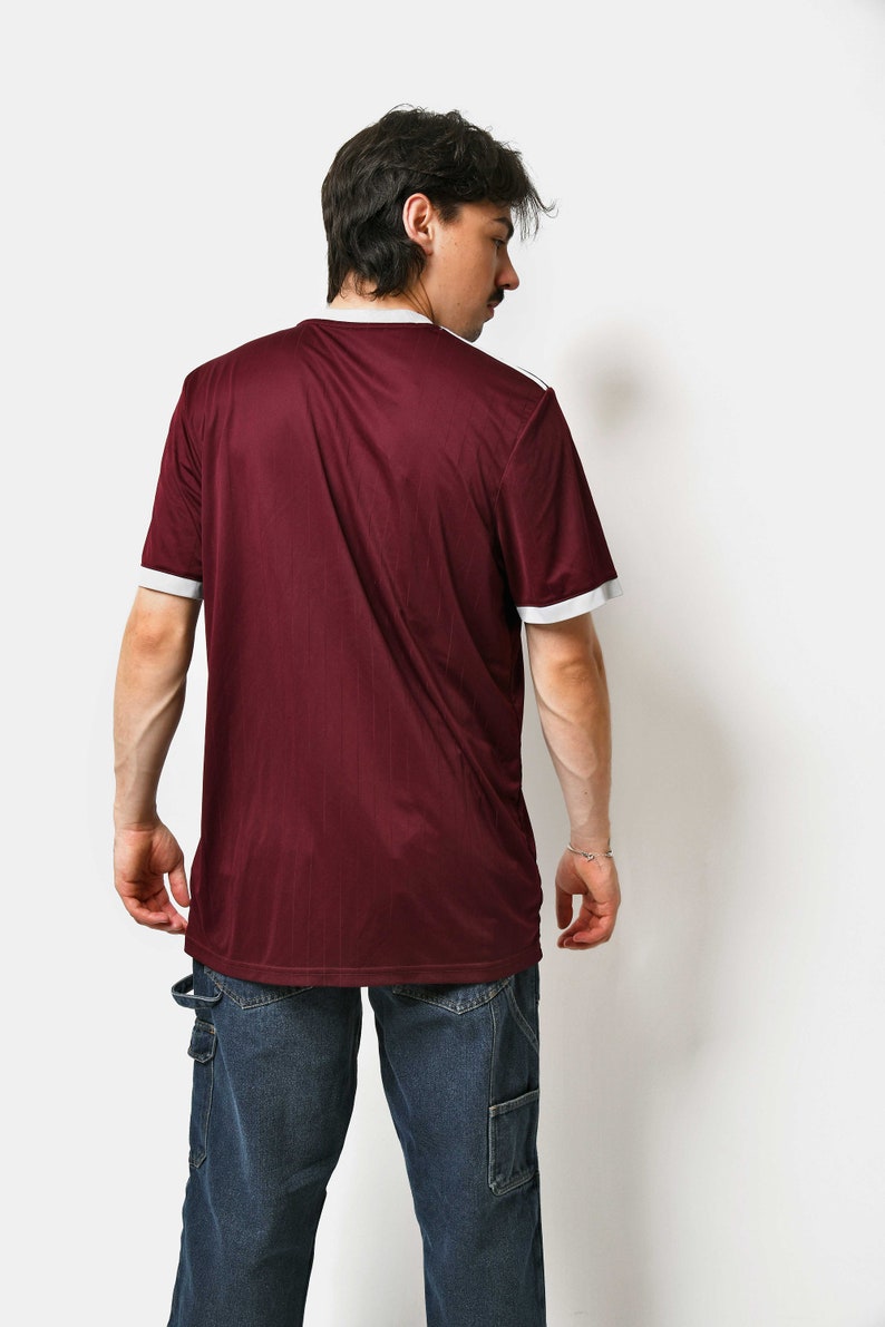 May include: A maroon t-shirt with white trim on the collar and sleeves. The t-shirt is made of a soft, comfortable material and has a relaxed fit. The person is wearing blue jeans.