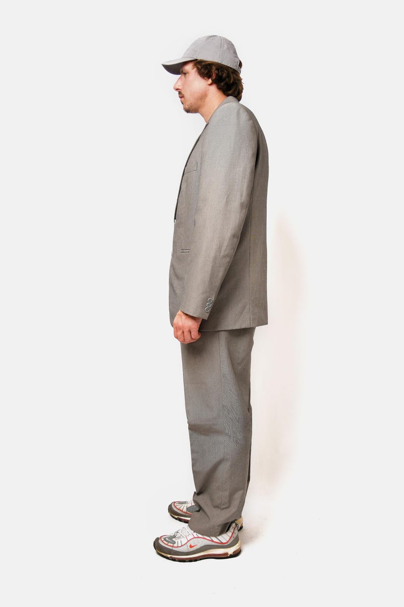May include: A gray suit with a matching gray cap. The suit jacket has a single button closure and the pants have a straight leg. The person is wearing white sneakers with red accents.