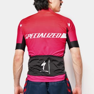 May include: A red and pink Specialized cycling jersey with black sleeves and white text. The jersey has a black waist pack with a Specialized logo. The person is wearing blue shorts with red stripes.