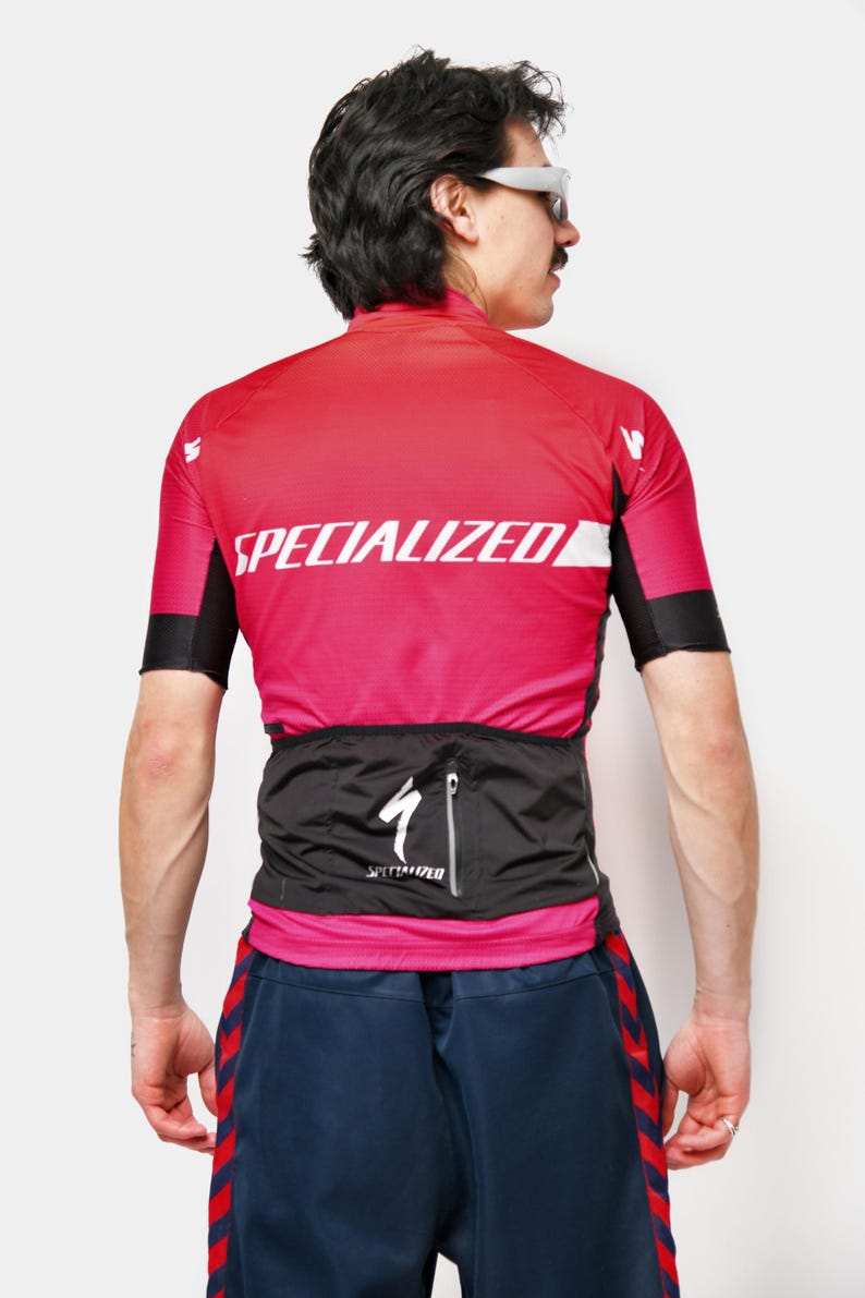 May include: A red and pink Specialized cycling jersey with black sleeves and white text. The jersey has a black waist pack with a Specialized logo. The person is wearing blue shorts with red stripes.