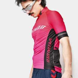 May include: A pink and black Specialized cycling jersey with the brand name prominently displayed. The jersey has a high collar and short sleeves. The person in the image is wearing silver sunglasses and navy blue athletic pants.