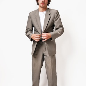 May include: A man wearing a gray suit with a gray cap and white sneakers. He is standing in front of a white background.