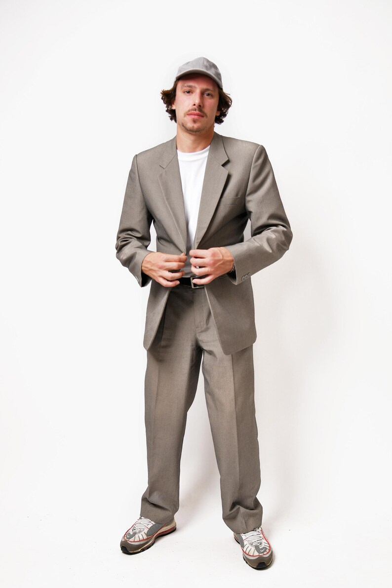 May include: A man wearing a gray suit with a gray cap and white sneakers. He is standing in front of a white background.