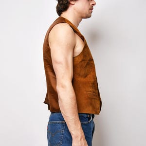 May include: A brown suede vest with a button closure. The vest is sleeveless and has a collar. The person wearing the vest is also wearing blue jeans and a brown baseball cap.