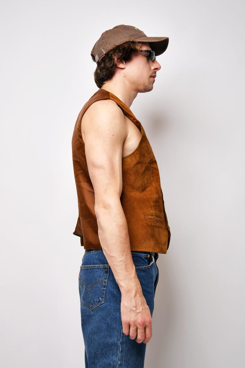 May include: A brown suede vest with a button closure. The vest is sleeveless and has a collar. The person wearing the vest is also wearing blue jeans and a brown baseball cap.