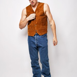 May include: A man wearing a brown suede vest, blue jeans, and brown sneakers. He is also wearing a brown baseball cap with the text "California University" on it and sunglasses.
