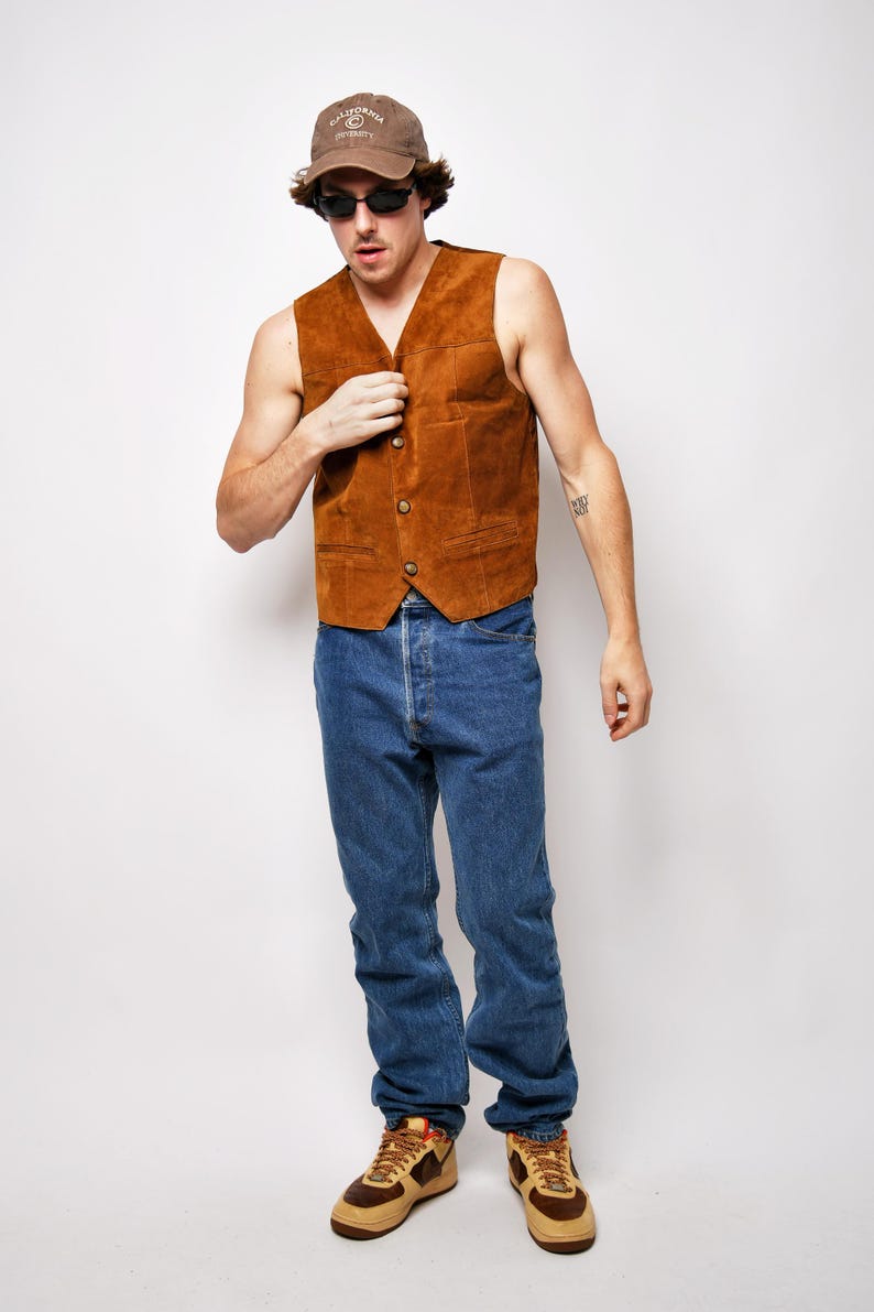 May include: A man wearing a brown suede vest, blue jeans, and brown sneakers. He is also wearing a brown baseball cap with the text "California University" on it and sunglasses.