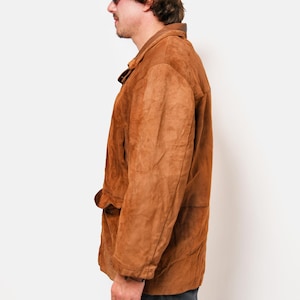 May include: A brown suede jacket with a button closure and a flap pocket on the left side. The jacket has a collar and long sleeves. The person is wearing a gray baseball cap.