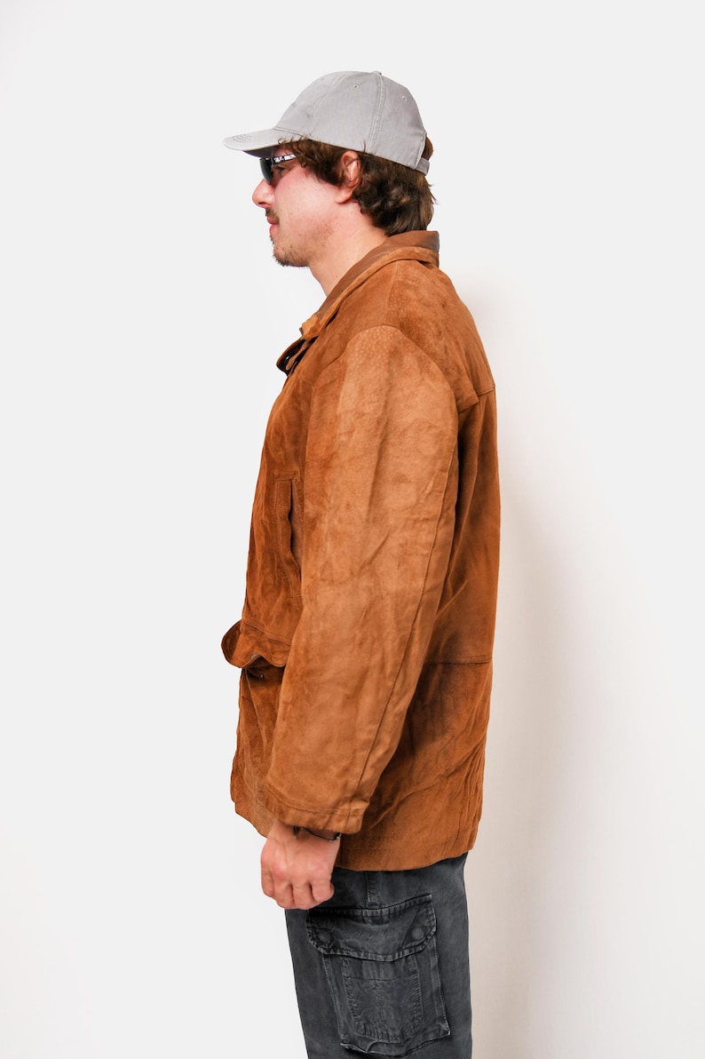 May include: A brown suede jacket with a button closure and a flap pocket on the left side. The jacket has a collar and long sleeves. The person is wearing a gray baseball cap.