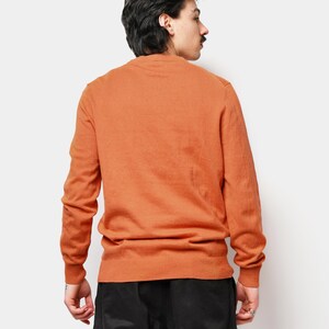 Puede incluir: Un su&eacute;ter de cuello redondo naranja quemado con mangas largas. El su&eacute;ter est&aacute; hecho de un material de punto suave y se usa con pantalones negros. El modelo est&aacute; de espaldas a la c&aacute;mara.