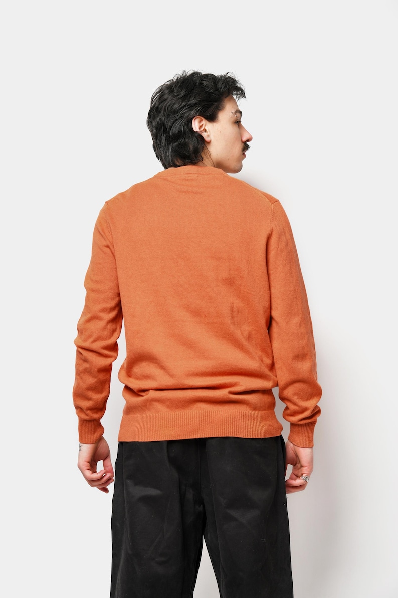 Puede incluir: Un su&eacute;ter de cuello redondo naranja quemado con mangas largas. El su&eacute;ter est&aacute; hecho de un material de punto suave y se usa con pantalones negros. El modelo est&aacute; de espaldas a la c&aacute;mara.