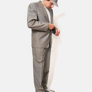 May include: A man wearing a gray suit, a gray baseball cap, and white sneakers. He is standing and looking down at his suit jacket.