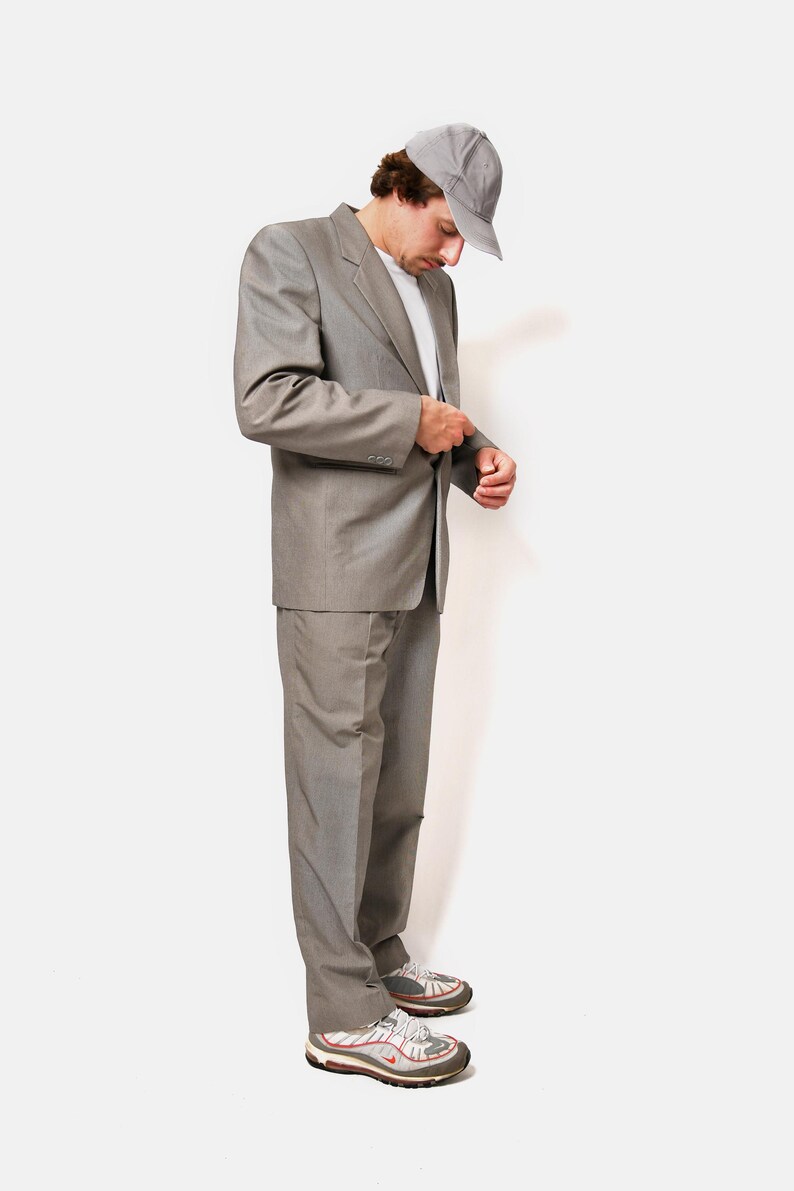 May include: A man wearing a gray suit, a gray baseball cap, and white sneakers. He is standing and looking down at his suit jacket.