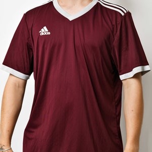 May include: A maroon short-sleeved t-shirt with white trim around the neck and sleeves. The shirt has a white Adidas logo on the left chest and three white stripes on the right shoulder.