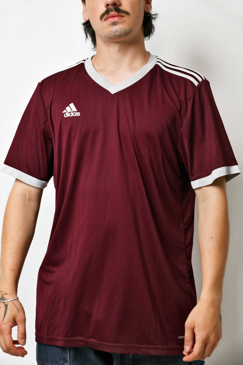 May include: A maroon short-sleeved t-shirt with white trim around the neck and sleeves. The shirt has a white Adidas logo on the left chest and three white stripes on the right shoulder.