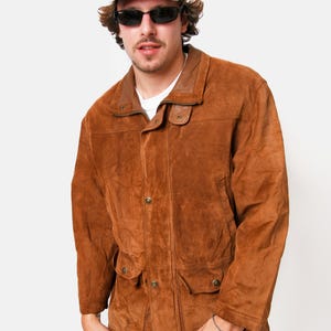 May include: A brown suede jacket with a collared neckline, button closure, and two flap pockets. The jacket has a light brown lining and a gray baseball cap is worn on top.