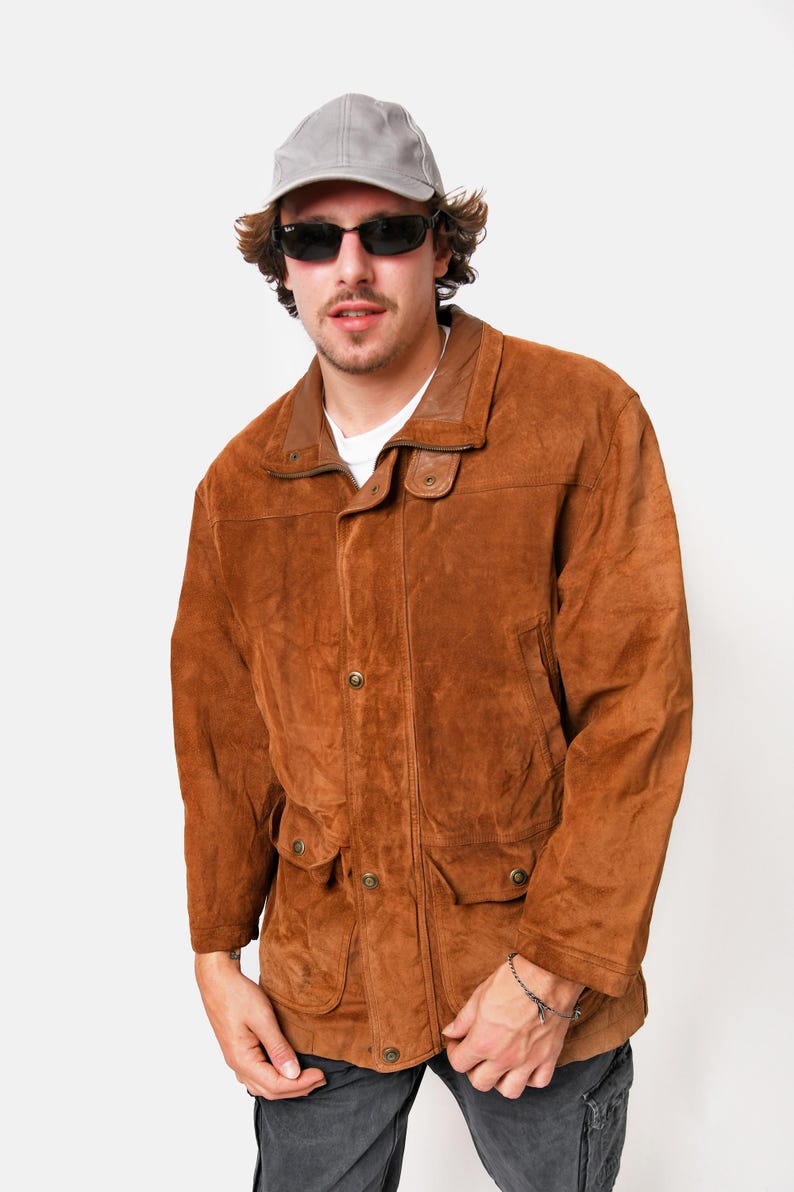 May include: A brown suede jacket with a collared neckline, button closure, and two flap pockets. The jacket has a light brown lining and a gray baseball cap is worn on top.