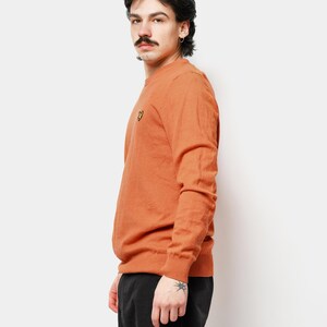 Puede incluir: Un su&eacute;ter naranja de cuello redondo con un peque&ntilde;o logotipo amarillo en el pecho. El su&eacute;ter est&aacute; hecho de un material suave y tiene mangas largas. La persona lleva pantalones negros.