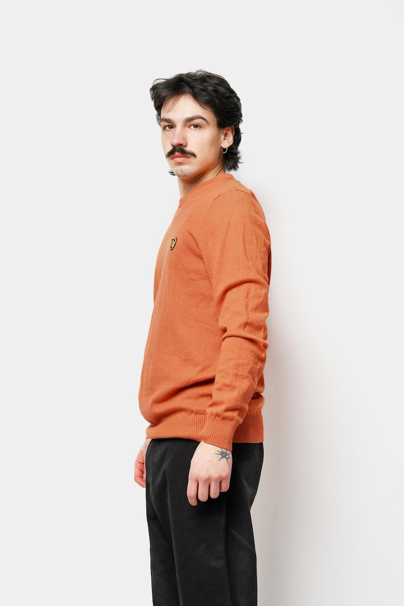 Puede incluir: Un su&eacute;ter naranja de cuello redondo con un peque&ntilde;o logotipo amarillo en el pecho. El su&eacute;ter est&aacute; hecho de un material suave y tiene mangas largas. La persona lleva pantalones negros.