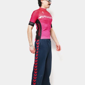 May include: A pink and black cycling jersey with the word "SPECIALIZED" printed on the front. The jersey is paired with navy blue track pants with a red chevron design down the side. The person is wearing sunglasses.