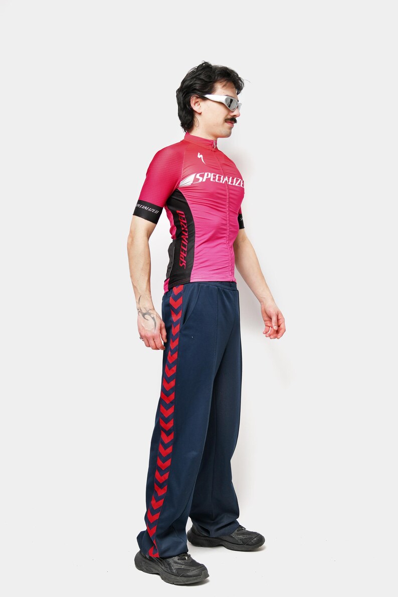 May include: A pink and black cycling jersey with the word "SPECIALIZED" printed on the front. The jersey is paired with navy blue track pants with a red chevron design down the side. The person is wearing sunglasses.