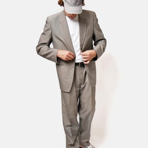 May include: A man wearing a gray suit jacket and matching pants, a white t-shirt, a gray baseball cap, and white sneakers.