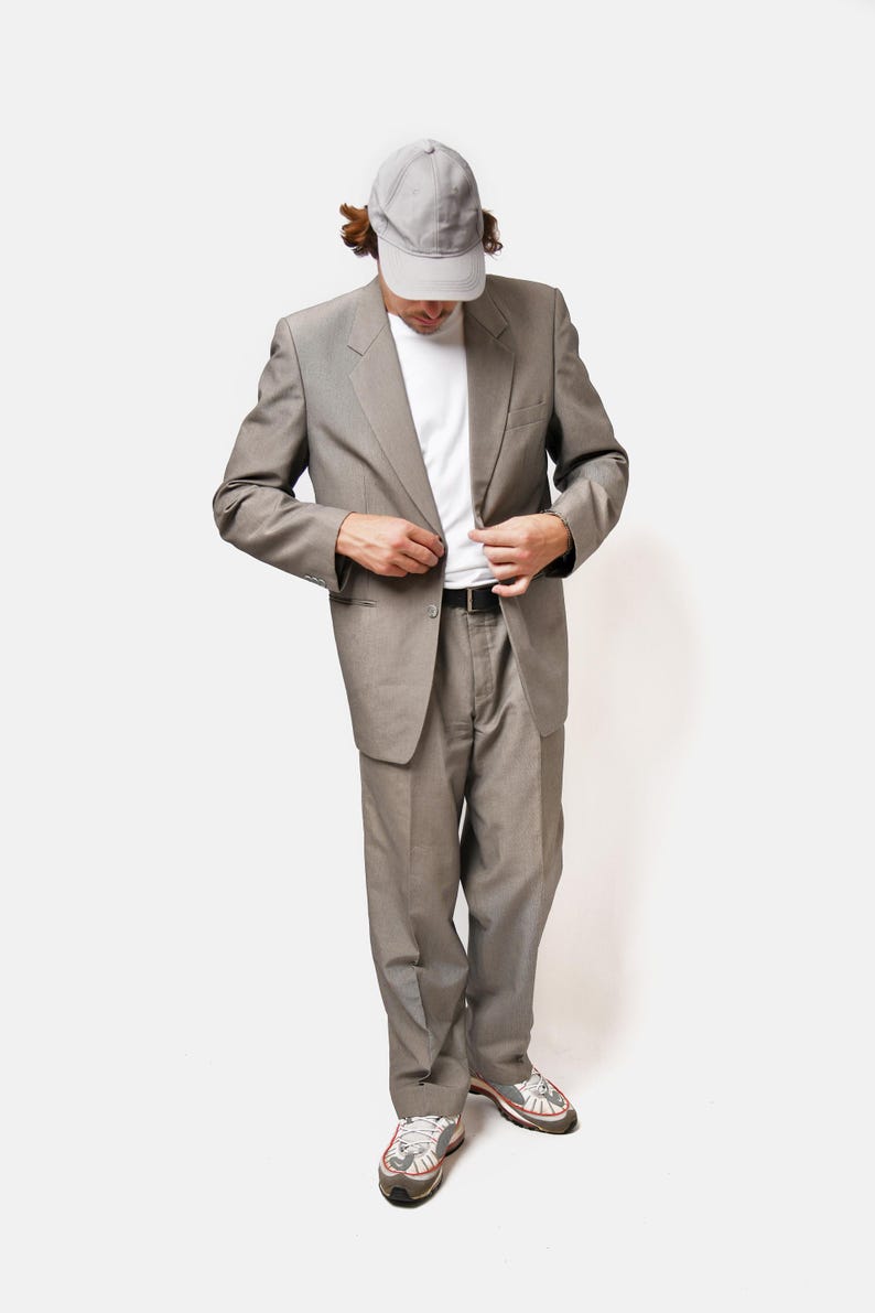 May include: A man wearing a gray suit jacket and matching pants, a white t-shirt, a gray baseball cap, and white sneakers.