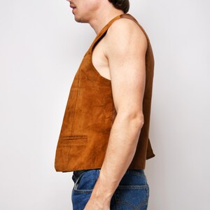 May include: A brown suede vest with a classic design. The vest has a pointed collar and a single button closure. The model is wearing the vest over a white shirt and blue jeans.