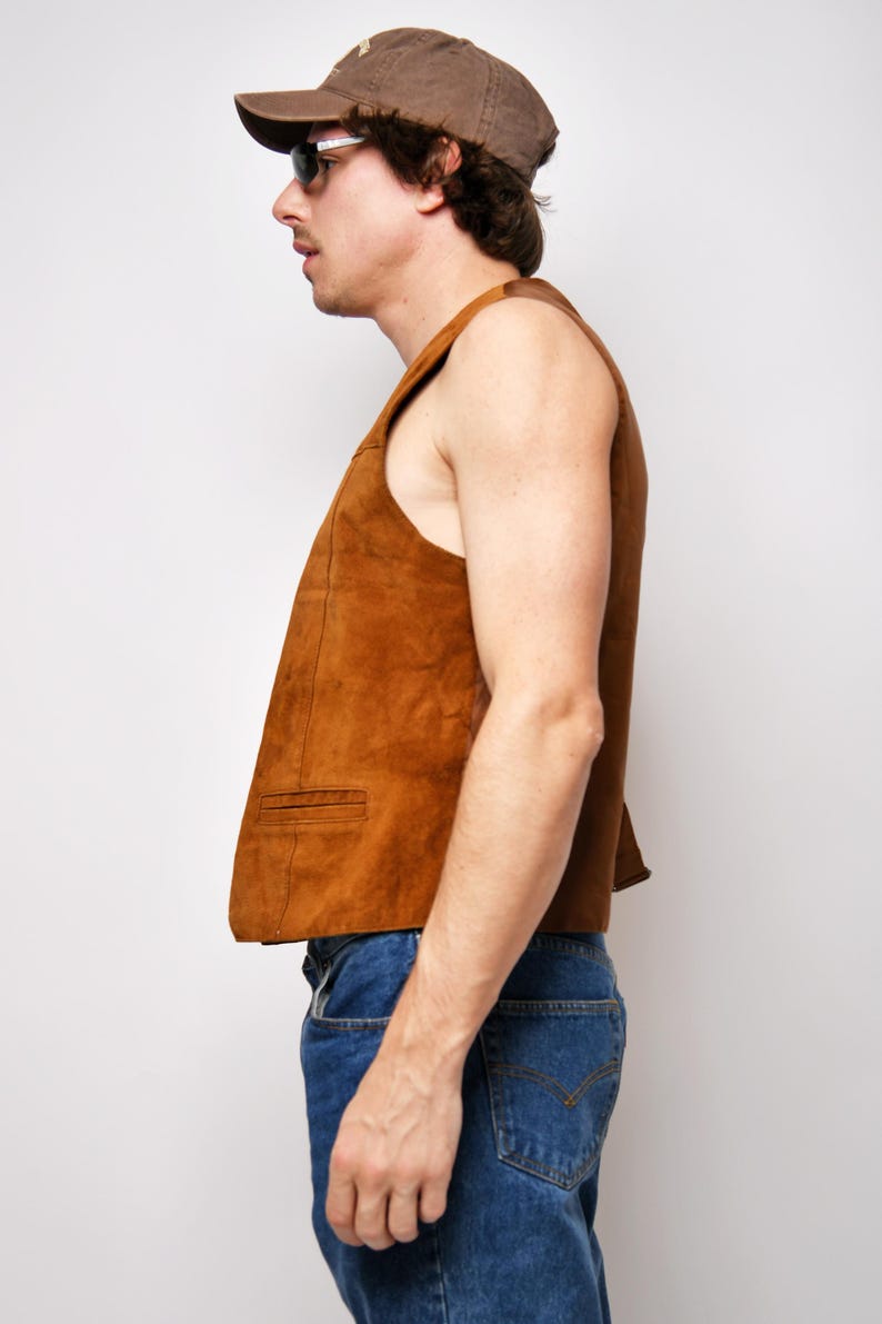 May include: A brown suede vest with a classic design. The vest has a pointed collar and a single button closure. The model is wearing the vest over a white shirt and blue jeans.