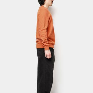 Puede incluir: Un su&eacute;ter naranja de cuello redondo y pantalones negros. El su&eacute;ter de manga larga es de color liso. Los pantalones son de color oscuro y tienen pernera recta. La persona lleva zapatillas negras.