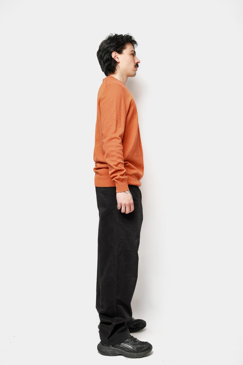 Puede incluir: Un su&eacute;ter naranja de cuello redondo y pantalones negros. El su&eacute;ter de manga larga es de color liso. Los pantalones son de color oscuro y tienen pernera recta. La persona lleva zapatillas negras.