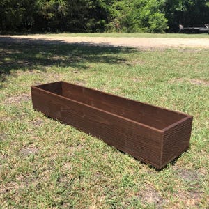 May include: A long, rectangular wooden planter box in a dark brown color. The planter is sitting on a grassy lawn.