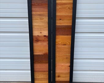 May include: Two wooden panels with a black frame. The panels are made of horizontal wooden planks in shades of brown and orange. The panels are leaning against a white garage door.