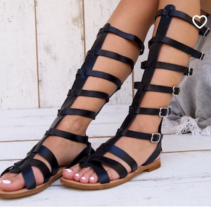 May include: Black leather gladiator sandals with multiple straps and buckles. The sandals have a flat sole and reach above the ankle.