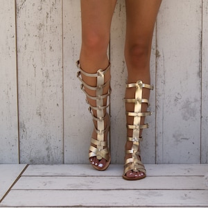 May include: A pair of gold gladiator sandals with straps up the leg. The sandals are on a white wooden surface.