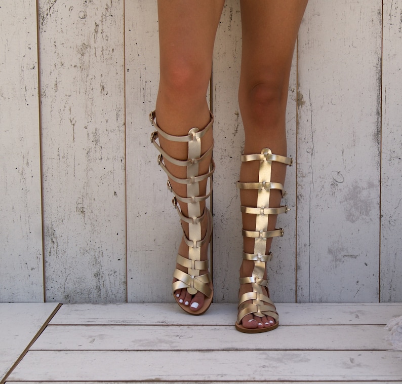 May include: A pair of gold gladiator sandals with straps up the leg. The sandals are on a white wooden surface.