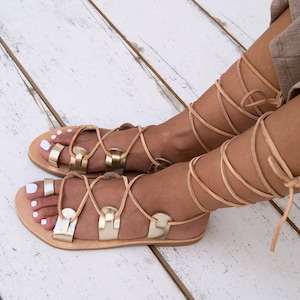 May include: Gold leather sandals with laces that tie up the leg. The sandals have a flat sole and are made of leather.