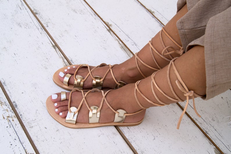 May include: Gold leather sandals with laces that tie up the leg. The sandals have a flat sole and are made of leather.