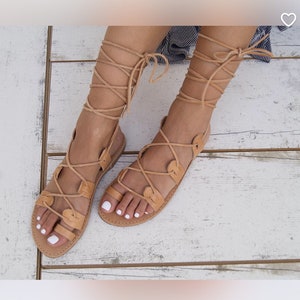 May include: Tan leather sandals with laces that tie up the leg. The sandals have a simple design with a rounded toe.
